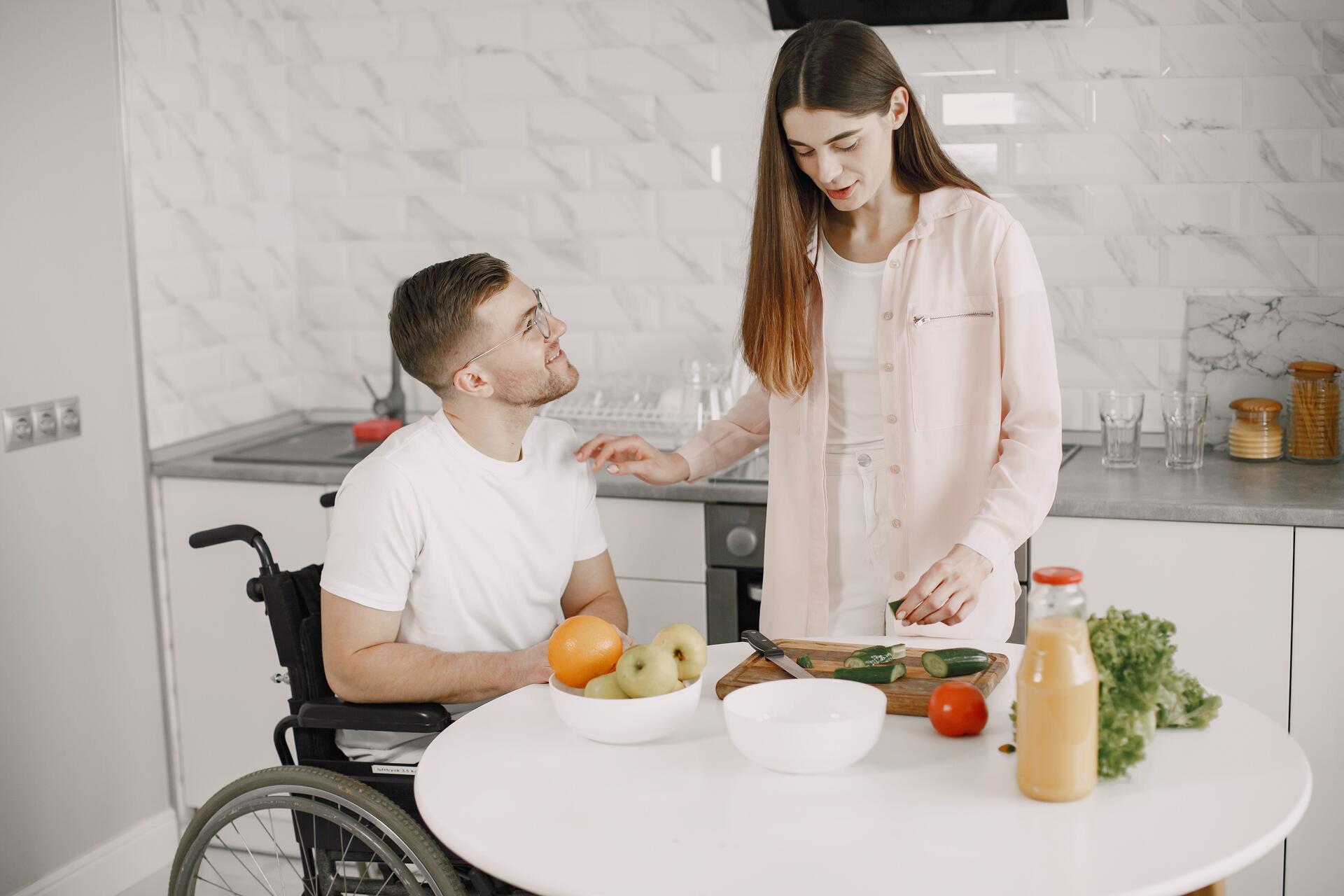 A man using a wheelchair shares a warm greeting with a woman outdoors near a white fence and greenery.