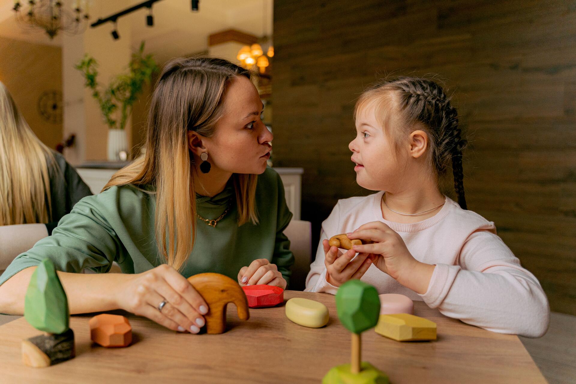 A woman and a young girl with Down syndrome sit together at a table, playing with wooden toys and talking closely.