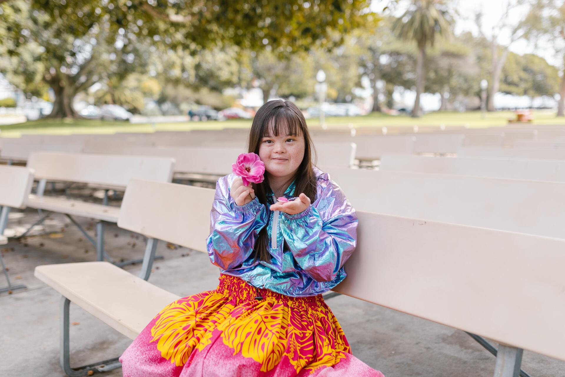 A young woman with Down syndrome smiles while holding a flower, sitting on a park bench outdoors.